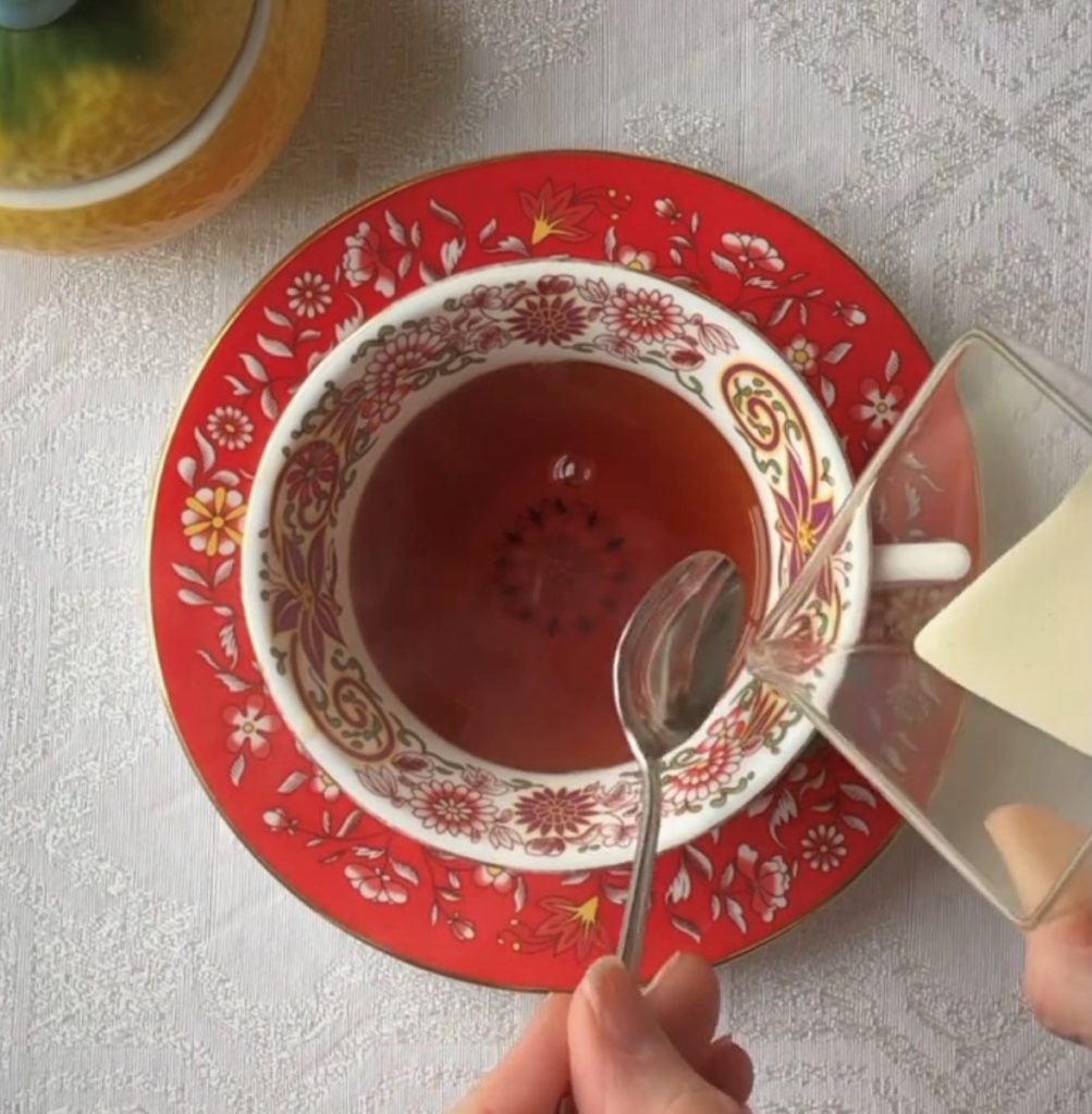 red and white Wedgwood teacup and saucer on a white tablecloth. A person is holding a spoon and about to pour in cream