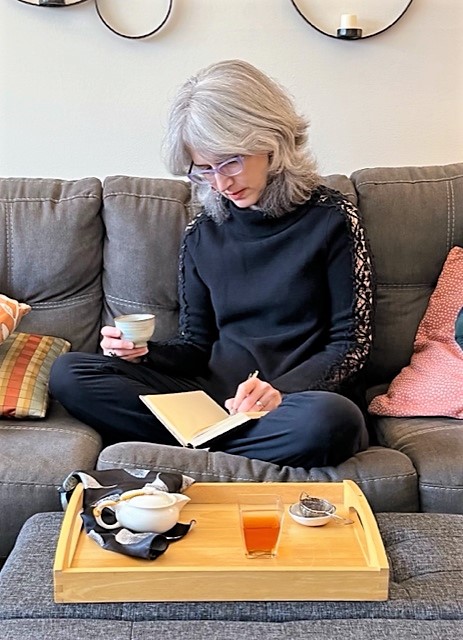 Image of Traci Levy (AKA Tea Infusiast) sitting on a sofa with a cup of tea in one hand and a notebook in the other.