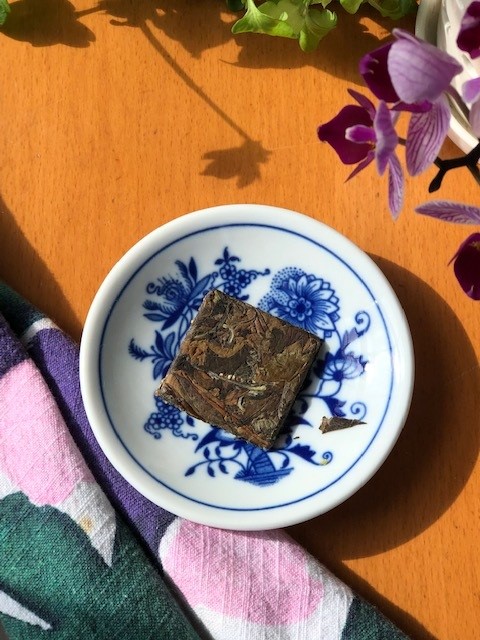 Square compressed tea cake with different shades of brown and green tea leaves on a blue and white dish sitting on a table beside a tea towel and a magenta orchid
