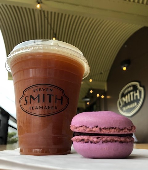 A tall cup of rich brown iced Nitro Chai with the Smith Teamaker logo on the cup, next to a purple macaron. The Smith Teamaker sign (black and cream) on the outside of the tasting room is in the background.