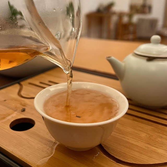 Inside T-Shop: pouring amber-colored Man Tang Xiang tea from a fairness pitcher to a teacup sitting on a tea tray next to an ivory teapot.