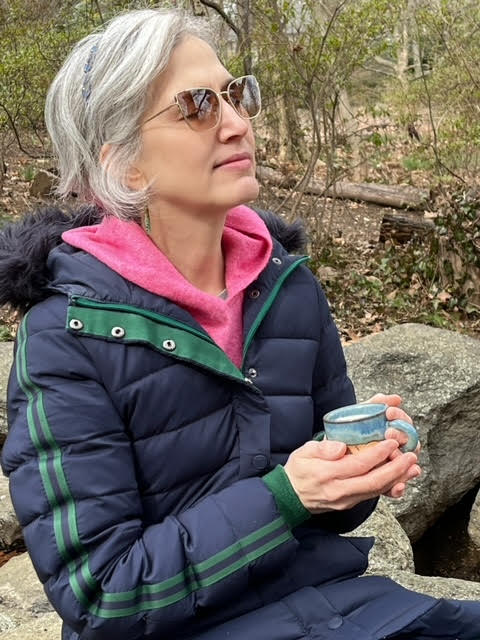 Traci Levy, a white woman with salt-and-pepper hair wearing a blue jacket and a pink hoodies and sunglasses, sitting outdoors and holding a cup of tea while resting.