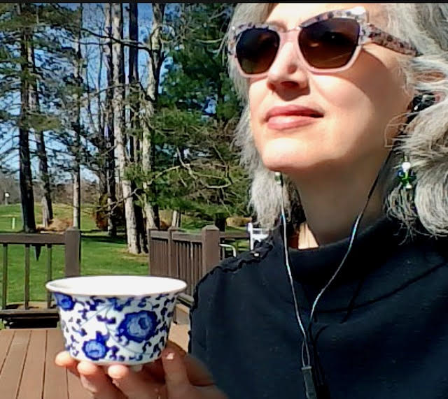 Traci Levy, a white woman with salt-and-pepper hair, wearing sunglasses and a black sweater, sitting outside and holding a blue and white teacup. Her face is serene and there are trees in the background.