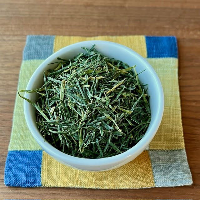 Barley Sprout herbal tea. Green leaves with touches of yellow  green, rolled to be long and thin. A cup of the leaves sits on a traditional Korean fabric coaster.