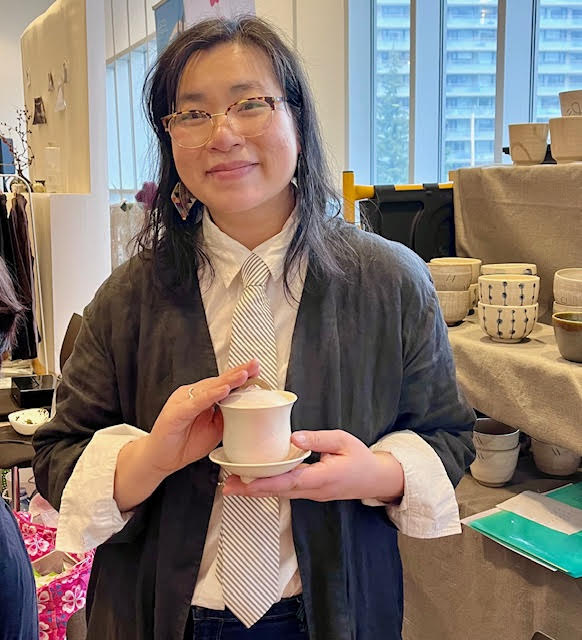 Helen of Secret Tea Time--a woman with dark hair and glasses wearing a white shirt with a striped black and white tie and black duster holding a cream and peach blush gaiwan she made.