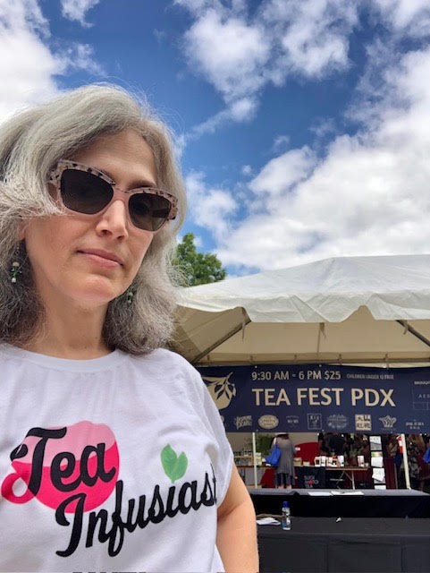 Traci Levy, AKA Tea Infusiast, a white woman whit salt and pepper hair wearing a t-shirt that says "Tea Infusiast" in front of a Teafest PDX sign.