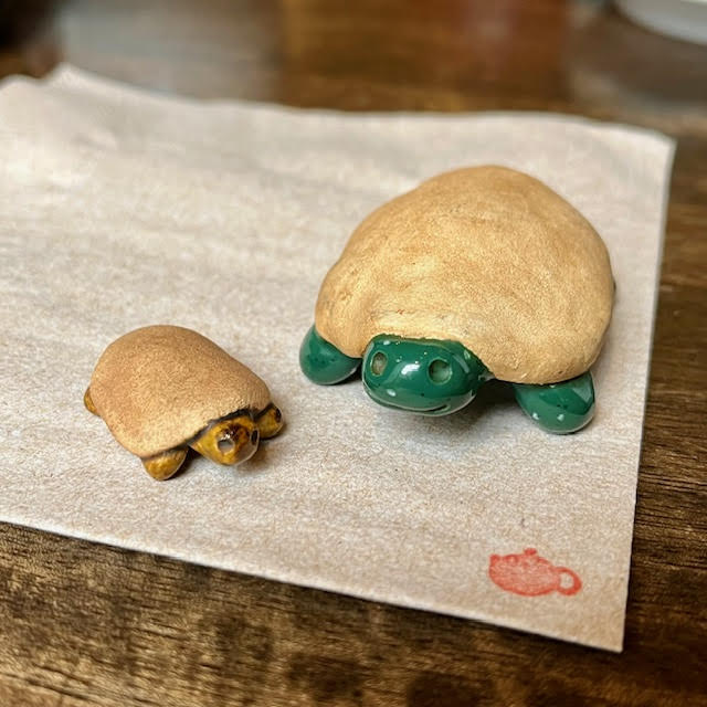 Two turtle tea pets--a tiny smiling clay turtle with a greenish brown face and feet and a larger smiling clay turtle with a bright green face and feet. Both have clay shells that have been darkened by pouring tea on them over time.