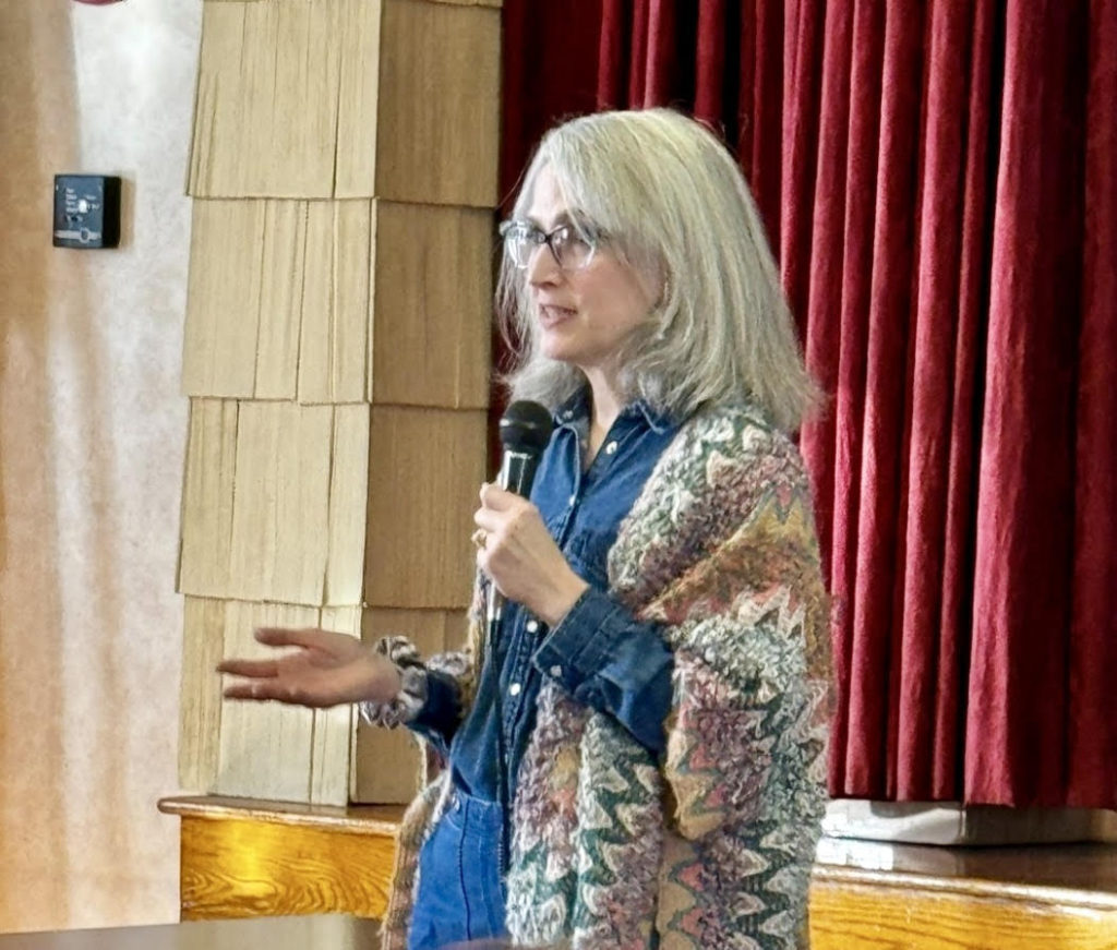 Traci Levy of Tea Infusiast--a white woman in her 50s with silver shoulder-length hair wearing glasses, a denim shirt and multicolor vest and holding a microphone will leading a tea event.