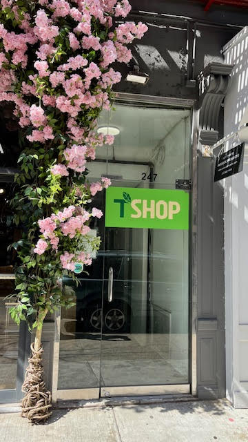 Exterior entrance to T Shop in Manhattan, New York. There are pink flowers with green leaves on the left side of the glass entrance door with a green sign that says "T Shop."