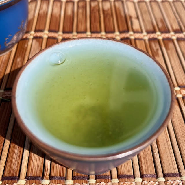 A green sencha tea in a handless blue cup with a brown rim. It sits on a bamboo mat.