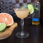 Grapefruit Bees Knees Mocktail in a class with a spring of rosemary and a lime garnish. A sliced grapefruit and lime are on a cutting board to the left.