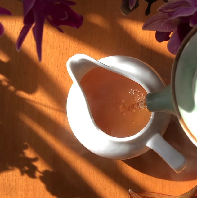 Green clay teapot pouring tea into a white ceramic fairness pitcher. Shadows of plants surround the teaware.