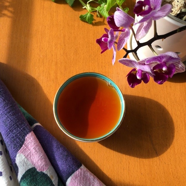 Tea the color of burnished copper in a teacup with a blue stripe at the top. The cup sits on a table next to a tea cloth and a magenta orchid.
