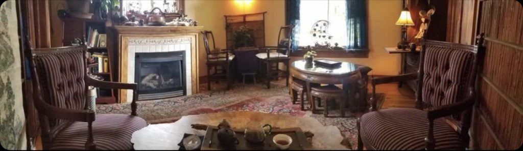 Wide-angle shot of one of the rooms inside Enthea Teahouse. There are cozy upholstered wooden chairs, tea tables with gongfu teaware, and a fire place. 