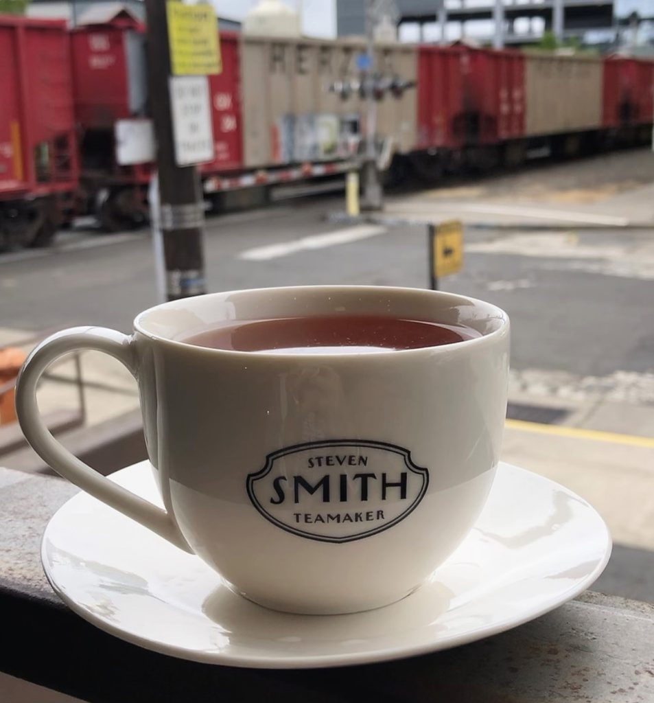 White ceramic cup and saucer with the black Smith Teamaker logo sitting on the rail of a porch. A colorful freight train is passing in the distance