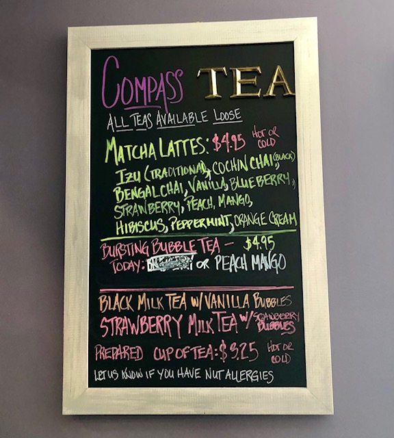 A framed chalk board. It says: "Compass Tea. All Teas Available Loose. Matcha Lattes: $4.95. Hot or Cold. Izu (traditinal, cochin chai, black), Bengali Chai, Vanilla, Blueberry, Strawberry, Peach Mango, Hibiscus, Peppermint, Orange Cream." "Busting Bubble Tea--$4.95." "Black Milk Tea w/ Vanilla Bubbles." "Strawberry Milk Tea w/strawberry bubbles." "Prepare Cup of Tea: $3.25. Hot or cold." "Let us know if you have nut allergies."