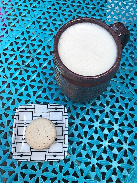 A white macaron with black specks of vanilla bean sits on a plate on a turquoise blue table next to a dark mug holding a creamy tea latte.