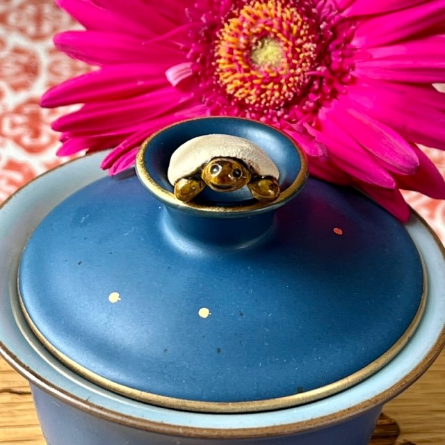 A tiny, smiling ceramic turtle teapet with unglazed shell perched on top of a blue and gold gaiwan. A magenta flower is in the background.