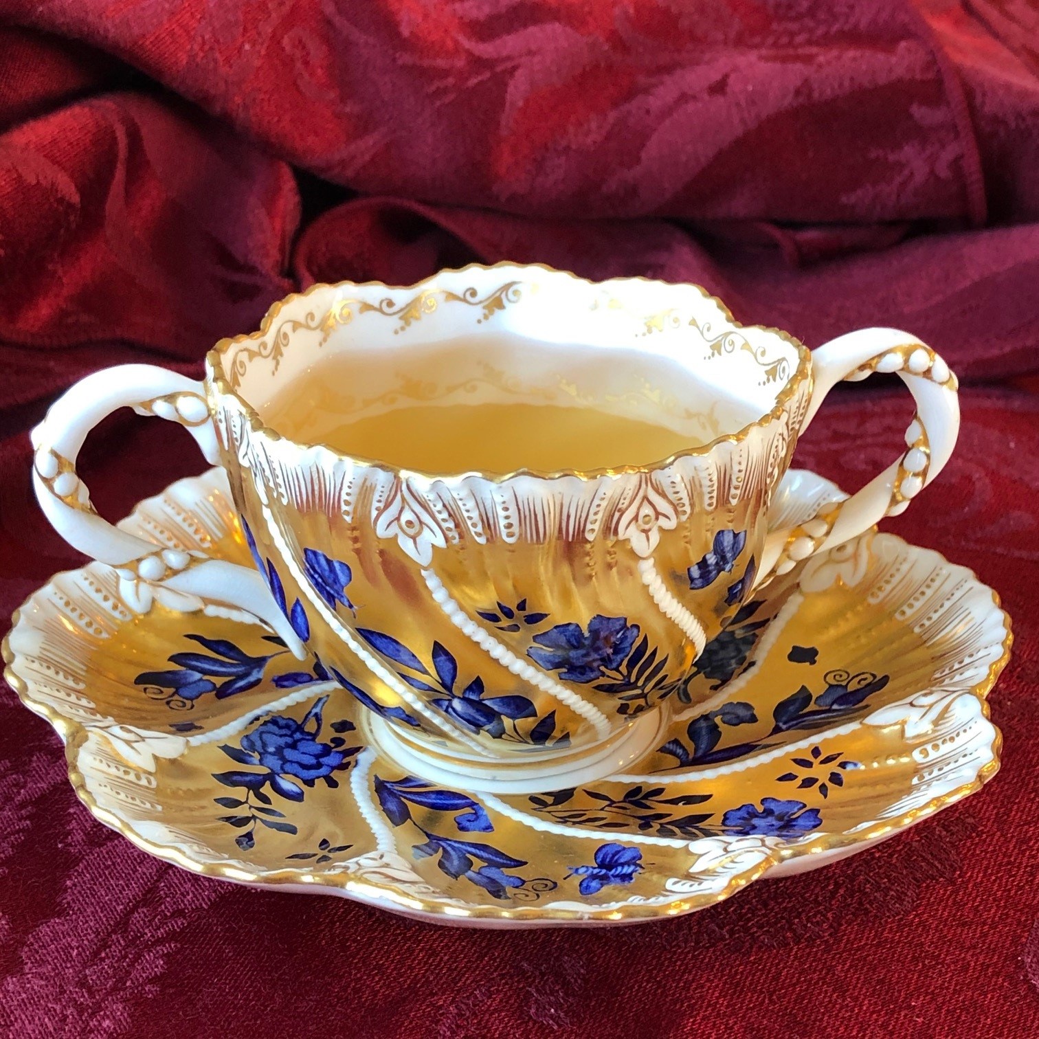 White and gold teacup with blue flowers on a crimson tablecloth. Traci's Sip & Listen story is about this teacup.