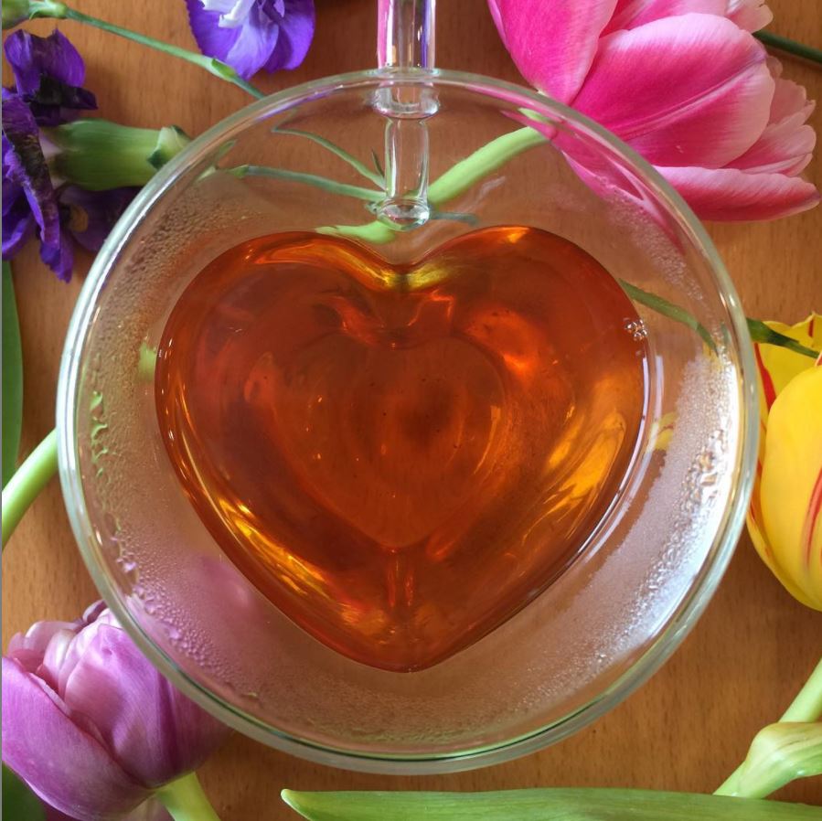 A glass, heart-shaped mug full of tea and love, surrounded by colorful tulips.