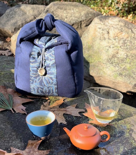 Handmade blue tea travel bag that taught me a lesson, on a rock with a clay teapot, glass fairness pitcher, and cup of tea.