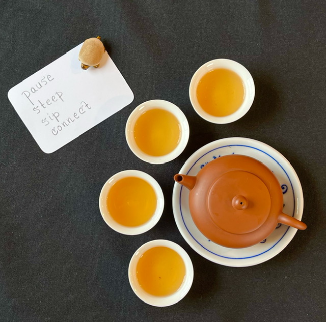 Four little cups of tea arrayed around a clay teapot. A teapet turtle sits on a card that says "Pause. Steep, Sip. Connect."