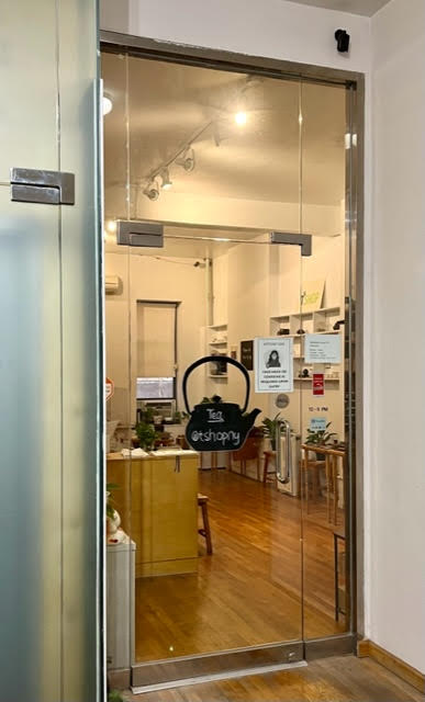 Looking through glass door of interior entrance to T-Shop, wooden floors and shelves, white walls, and lovely teaware.