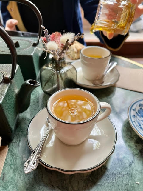 Inside Paquita: Two white cups with blue rims and matching saucers sitting on a deep green table with a iron teapots and a small bouquet of dried flowers. One person is in the process of pouring some honey into one of the cups of tea.