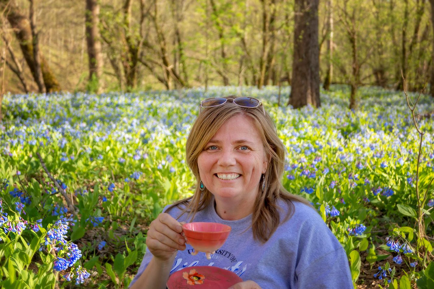 Tea on the Trail's Nicole McKinney--a blond woman, smiling, with sunglasses on top of her head, holding a footed pink teacup and matching saucer, in front of a field of bluebells.