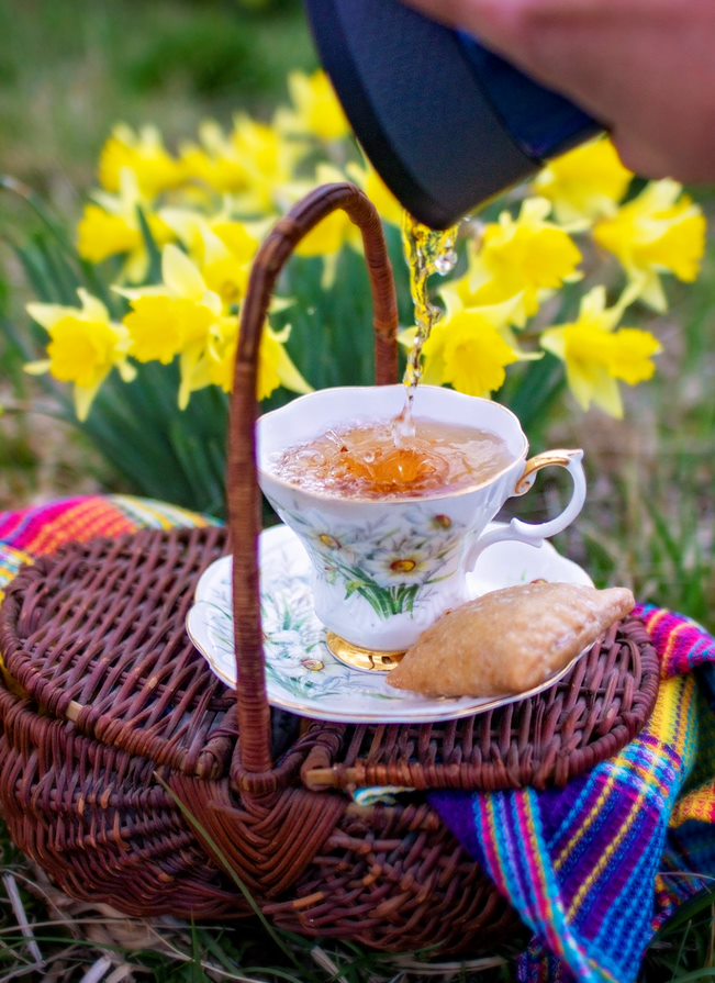 Tea on the Trail's photo of a white teacup with flowers on a saucer on top of a wicker basket with daffodils in the background. 