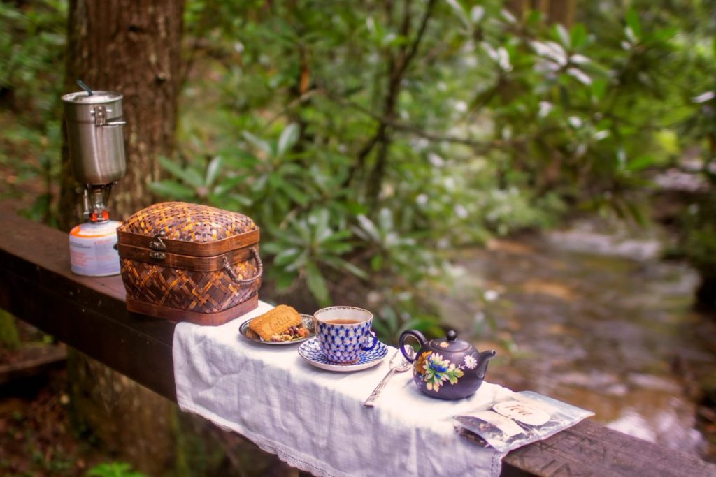 Tea on the Trail photo of a pocket stove, wicker basket, pastry, cup and saucer full of tea, and brown flowered teapot on the railing of a bridge over a stream.