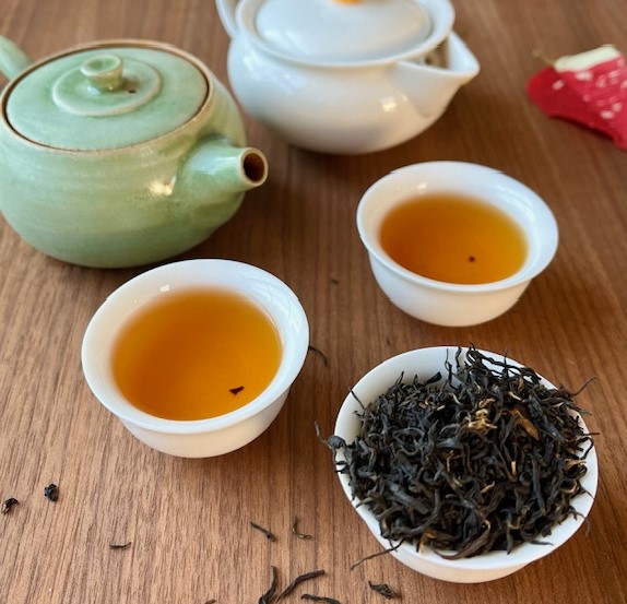 Two cups of Huiming hong cha tea next to a cup of the dried tea leaves and a white porcelain and a green clay teapot.