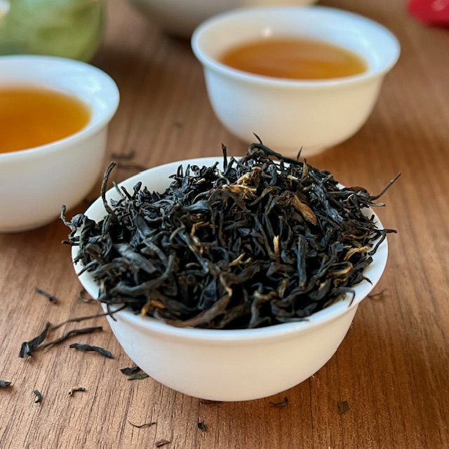 A close up of deep brown and caramel tea leaves of Huiming hong cha. Two cups of the steeped tea are in the background.