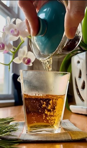 Tea Infusiast pouring Guan Yin Hong tea from a gaiwan into a fairness pitcher. The tea shows bubbles from the force of a pour. A white and magenta orchid is in the background.