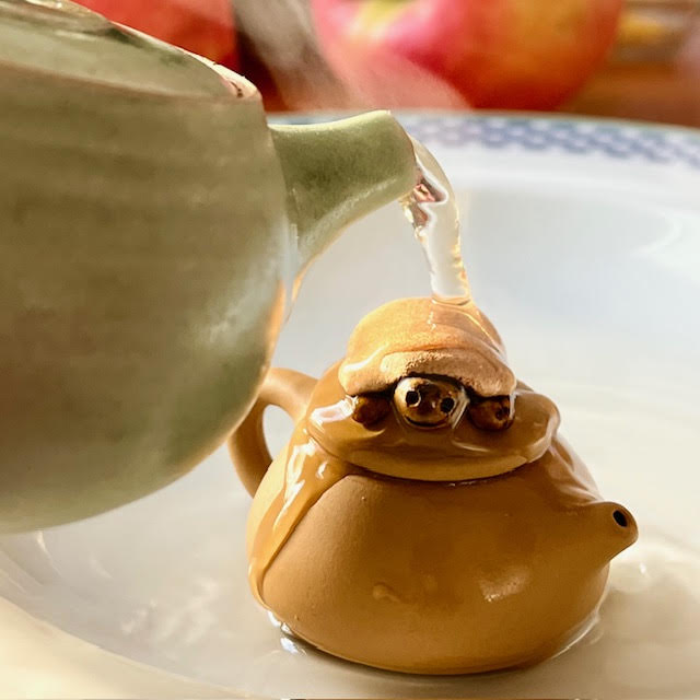 A green clay teapot pouring tea over a small clay turtle teapot on top of a tiny clay teapot.