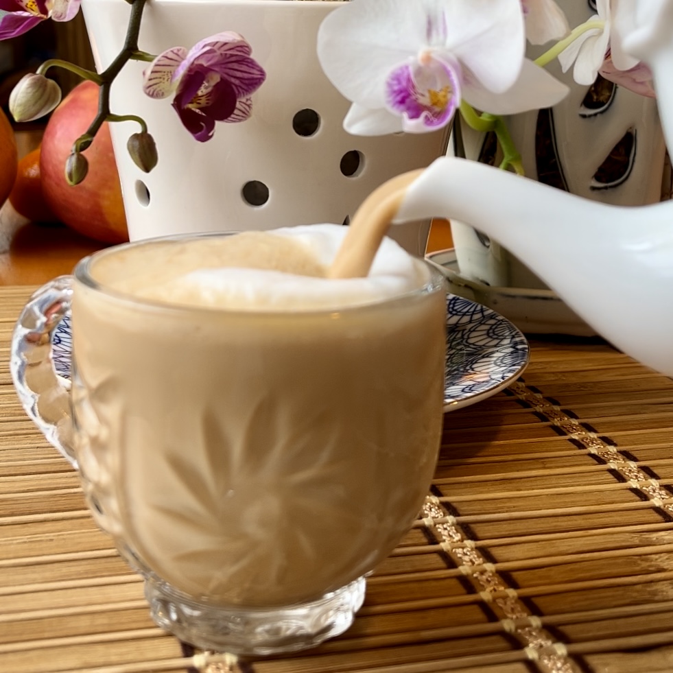 The spout of a white tea kettle pours masala chai into a glass mug. There are orchids in the background. An image to represent filing our cups and helping other women fill their cups.