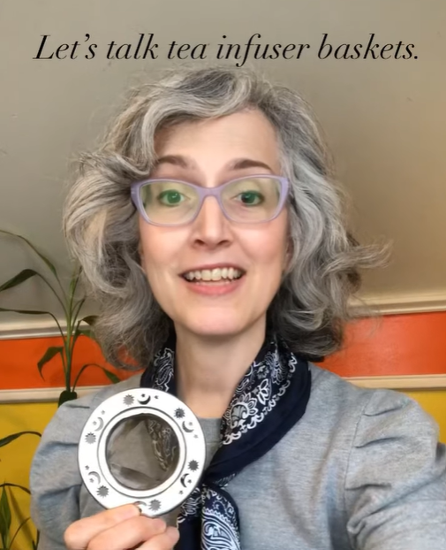 Photo of Traci Levy of Tea Infusiast --a white woman in her 50s wearing lavender glasses, a gray shirt, and a blue and white scarf around her neck holding a tea infuser.