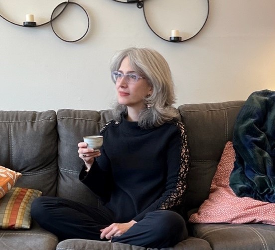 Photo of Traci Levy, writer of Tea Infusiast News, holding a teacup while seated on a sofa. Traci is a white woman with silver hair wearing violet glasses and a black sweater and pants.