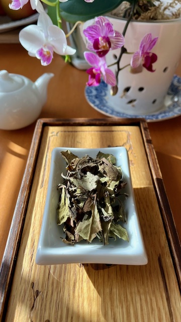 Tipsy Garden Barrel #2 tea leaves--flat, slightly broken greenish-brown leaves--in a white rectangular dish on a tea tray. A small porcelain teapot and blooming white and fuchsia orchids are in the background.