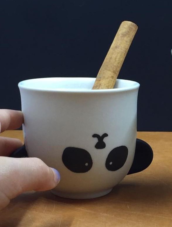 Ceramic teacup with the upside down face of a panda bear. A small hand is reaching for the cup. A cinnamon stick is visible poking out of the cup.