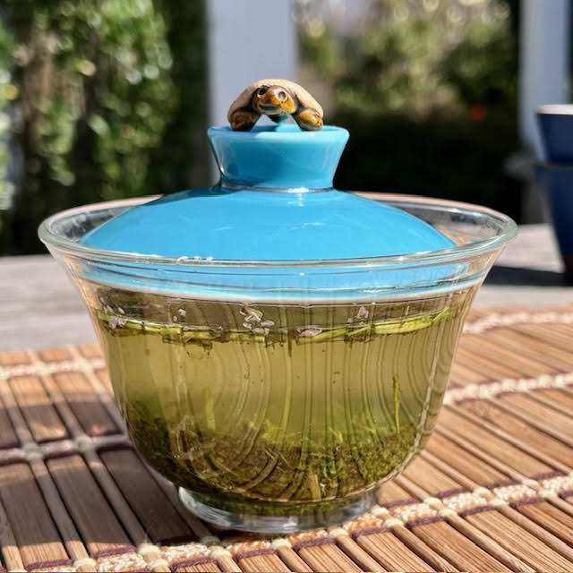 Sakura Kukicha creating a vibrant yellow green tea in a glass gaiwan with a turquois lid. A turtle teapet is on top of the gaiwan lid.