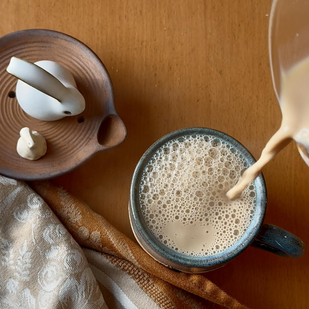 Pouring masala chai into a mug so that tea-bubbles form. Nearby is a brown cloth napkins and a clay tray with two white rabbit tea pets.