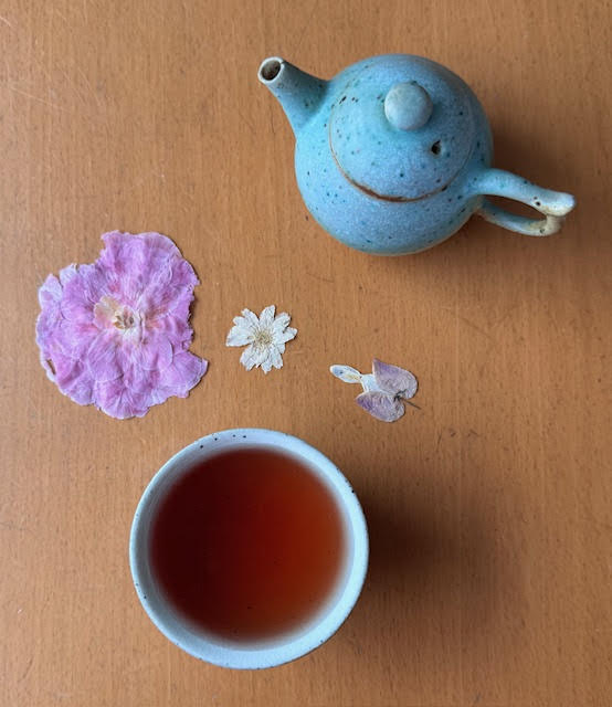 Dark amber-red steeped Phoenix Dan Cong in a small white teacup without a handle on a table next to a glazed blue clay teapot and dried flower petals