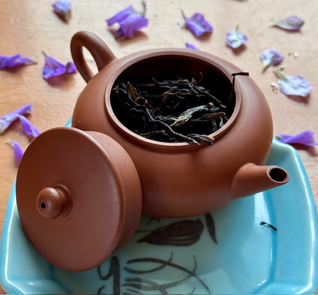 Red clay teapot with wiry dark brown tea leaves sticking out the top. Flower petals are in the background.