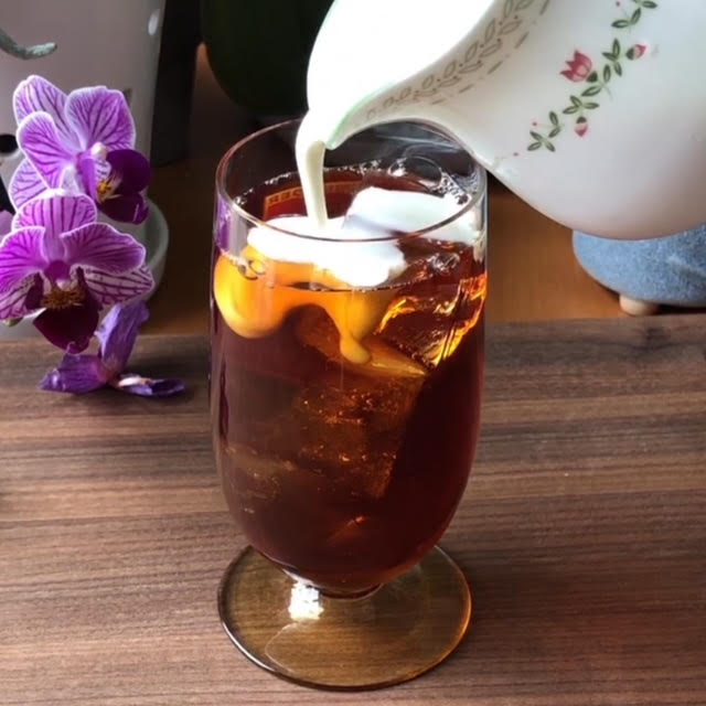 Pouring cream from a white creamer with a pink and green flower into a tall glass of black iced tea