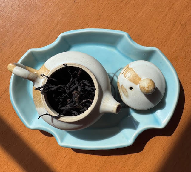 Jiang Hua tea leaves peeking out of a glazed white and tan teapot on a sky blue ceramic tray