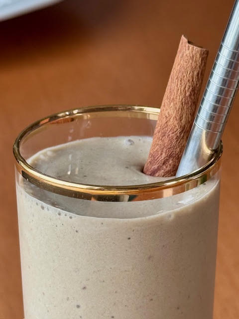 Close up of a Hojicha Banana Smoothie in a glass with a gold rim, a silver metal straw and a cinnamon stick garnish