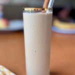 Hojicha Banana Smoothie in a tall glass with a gold rim. A silver metal straw and a cinnamon stick peek out of the glass