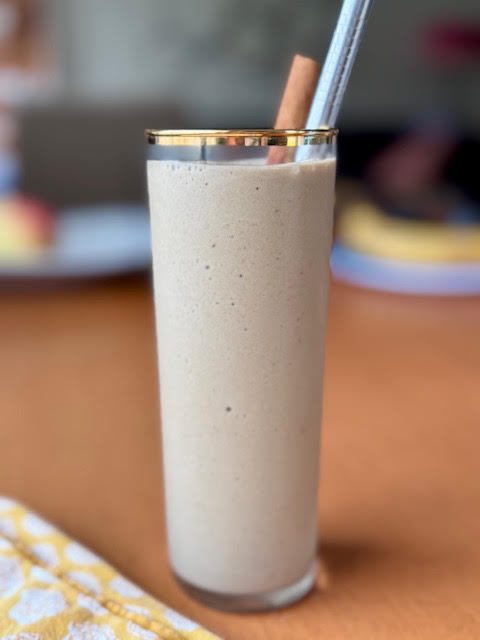 Hojicha Banana Smoothie in a tall glass with a gold rim. A silver metal straw and a cinnamon stick peek out of the glass