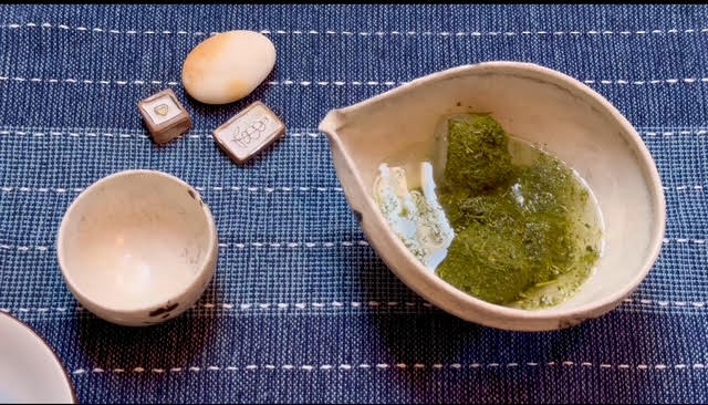 koridashi, or ice-brewed tea--green tea leaves over ice in a spouted bowl next to a white cup, a pebble, and two decorative ceramic tiles